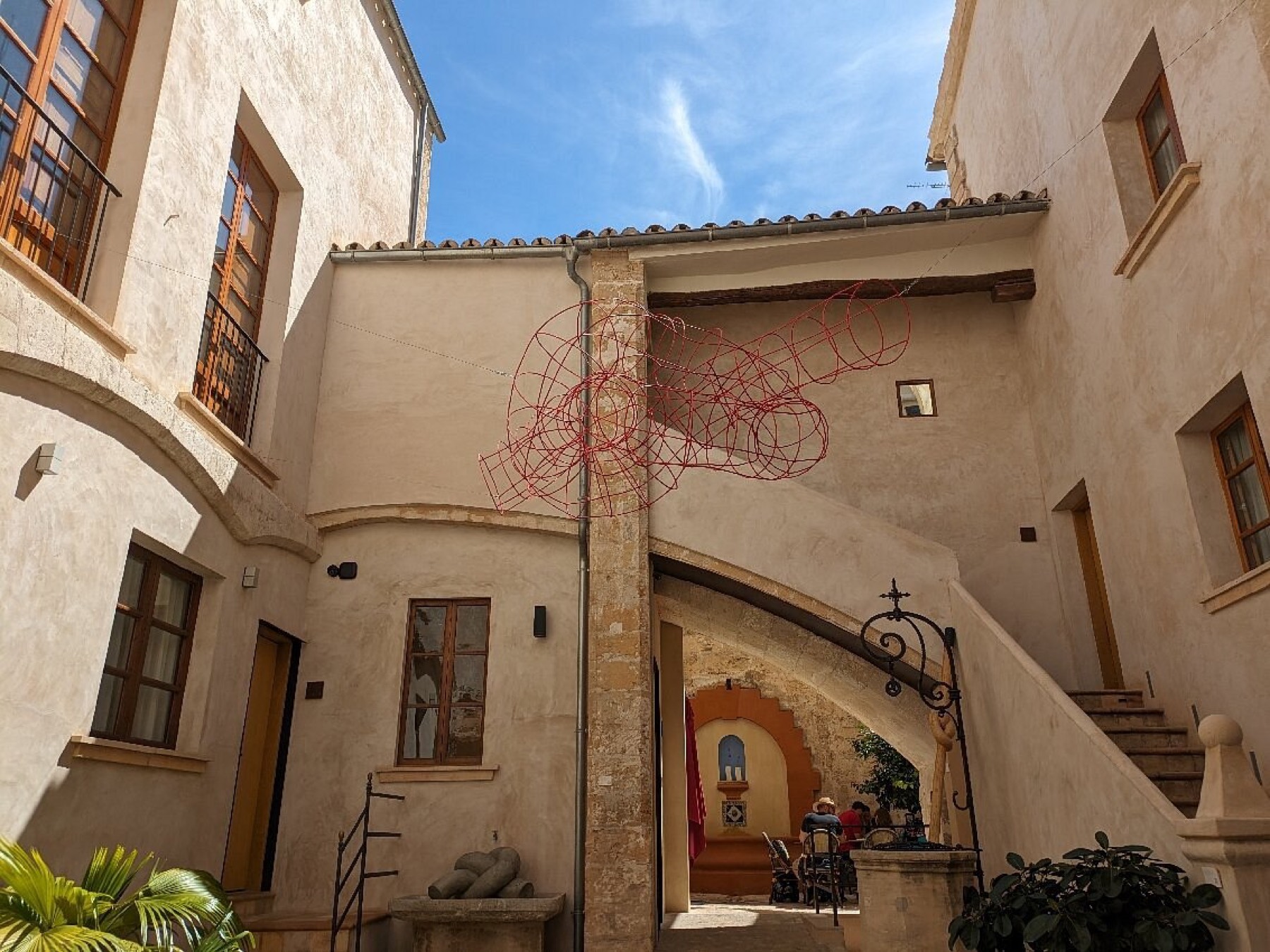 The courtyard of Can Cirera by day: a tall pointed stone archway, an external staircase, ochre walls and a stone well-head under a deep blue sky