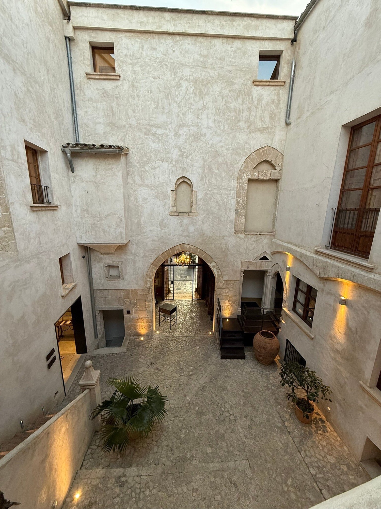 The stone courtyard of Hotel Can Cirera at golden hour, looking up at pointed Gothic windows and a warmly lit archway over a paved patio with a terracotta amphora