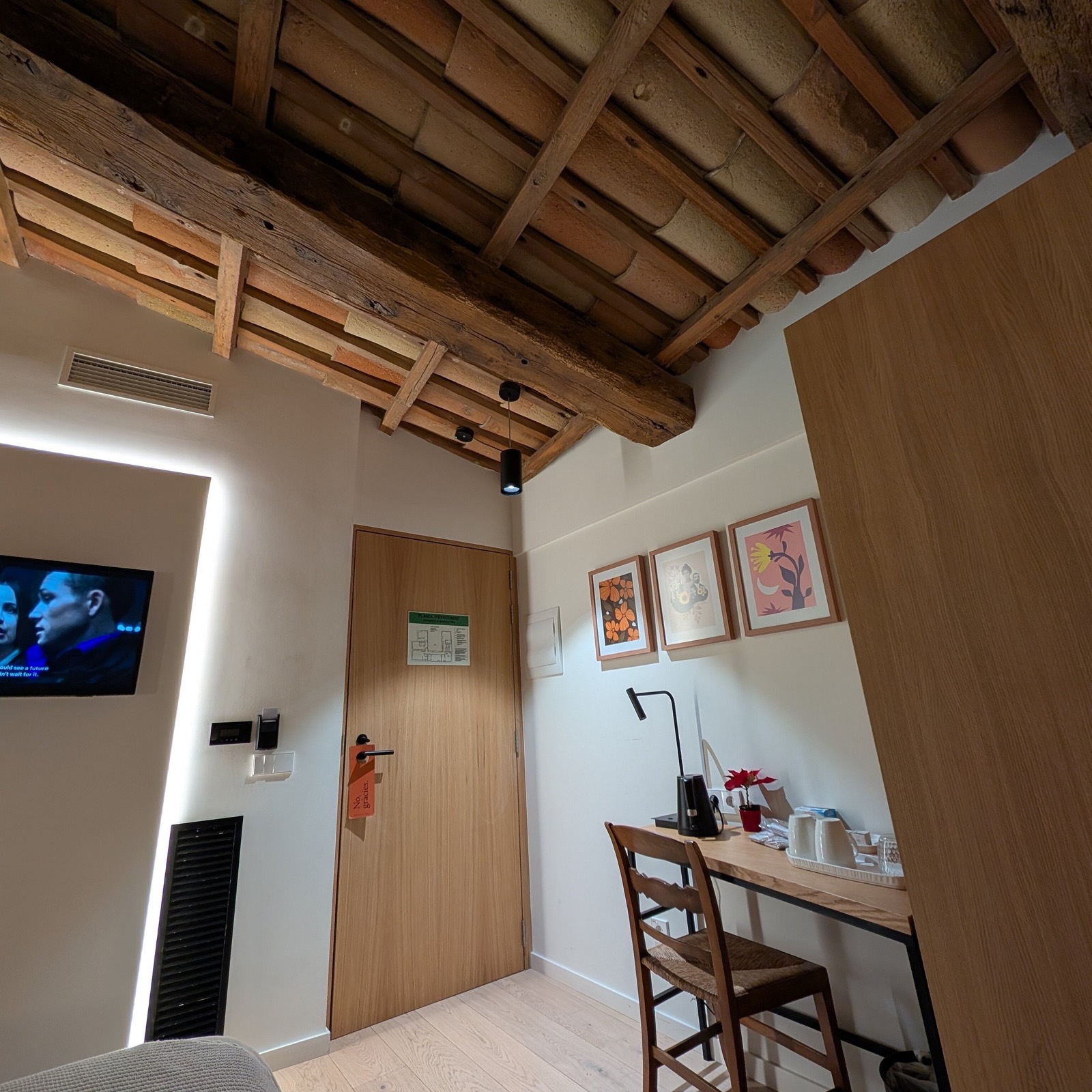 A Can Cirera room with an exposed timber-and-terracotta beamed ceiling, white walls, framed prints, oak joinery and a small writing desk