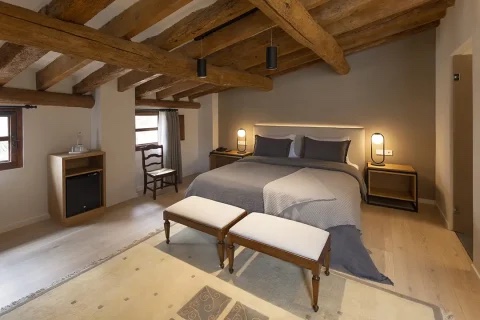 A suite room with exposed original timber ceiling beams, a king bed with grey linen, twin bedside lamps and a small writing desk by the window