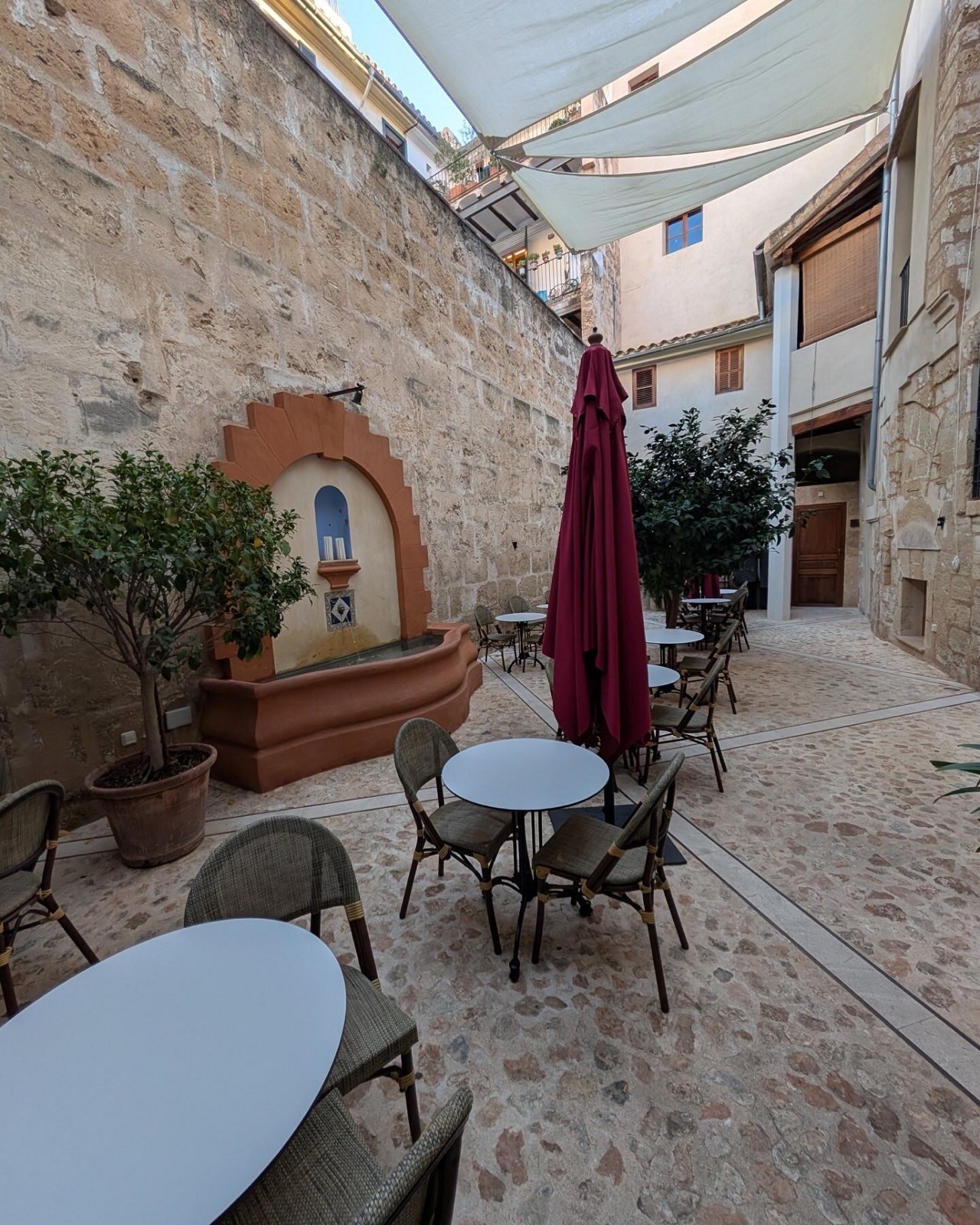 The courtyard set for the afternoon: bistro tables under a pale shade sail between stone walls, a small wall fountain and potted bay trees